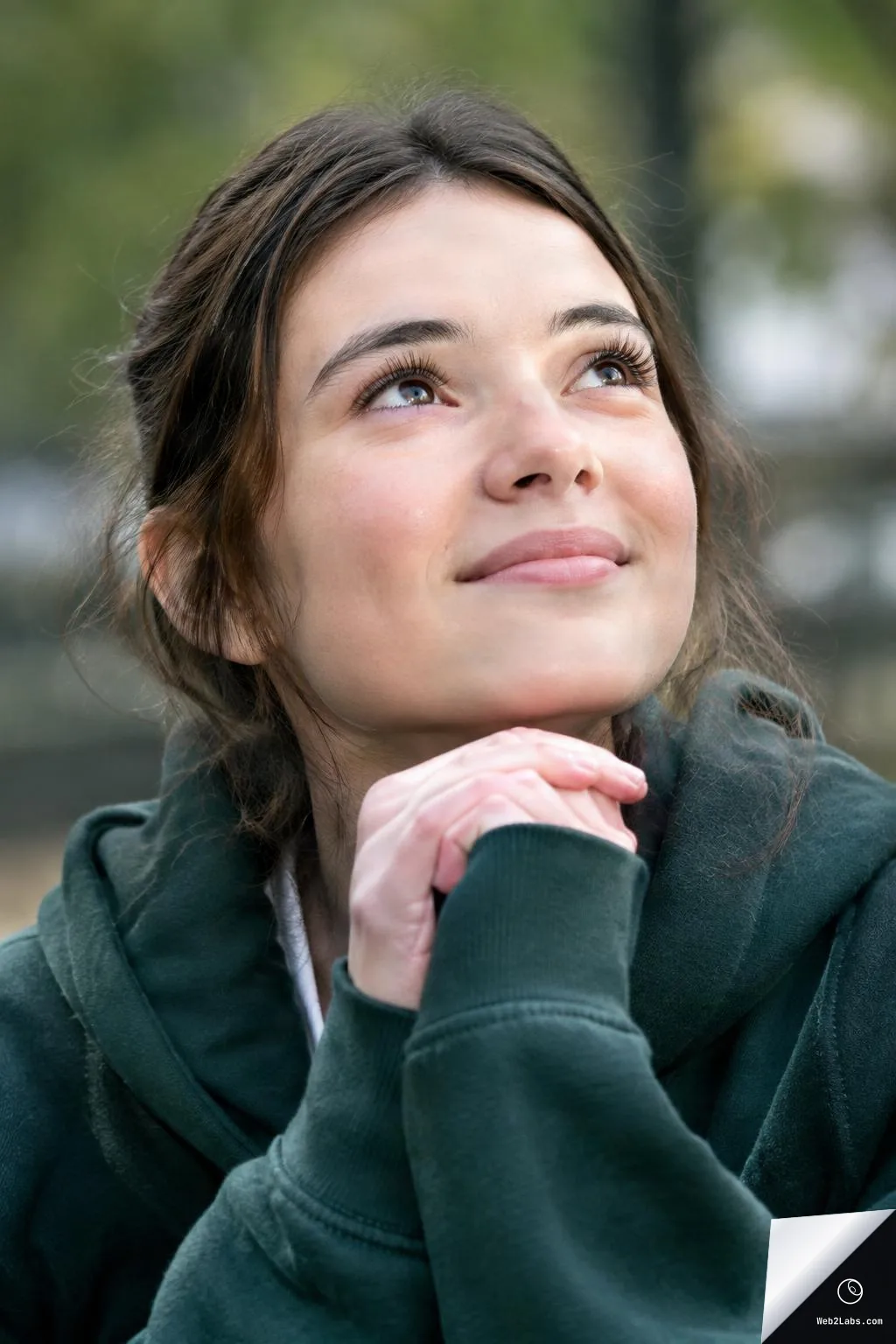 Close-up of a young woman in a dark green hoodie with hands clasped under her chin, looking upward with a gentle smile against a blurred outdoor background.