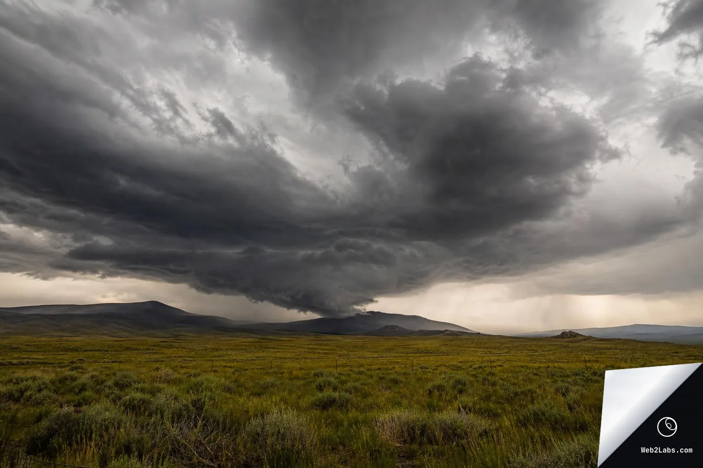 Dark, layered storm clouds looming over a wide green grassland with low hills on the horizon, photographed from near ground level.