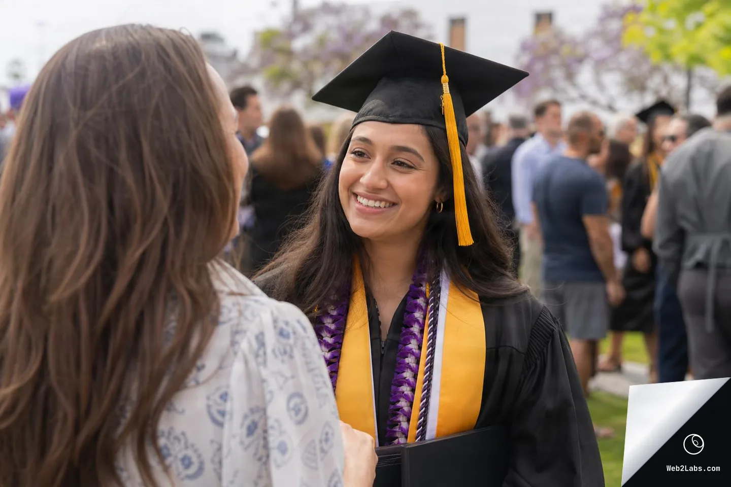 Over-the-shoulder view of a woman speaking with a smiling graduate wearing a black cap and gown with a gold stole and purple lei at an outdoor graduation gathering, with other attendees blurred in the background.