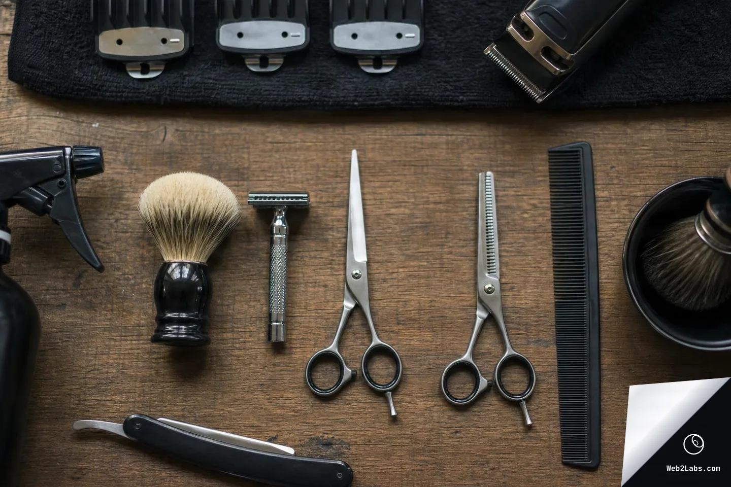Overhead view of barber tools on a wooden table, including scissors, thinning shears, comb, safety razor, straight razor, shaving brush, spray bottle, clipper guards, and an electric trimmer partially visible at the top right.