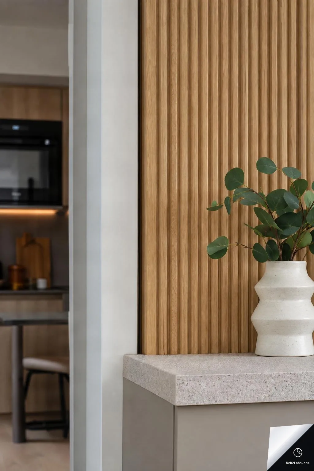 Vertical photo of a modern interior corner with a warm slatted wood wall, a light stone countertop edge, and a white ceramic vase holding green leafy stems; a blurred kitchenette is visible in the background.