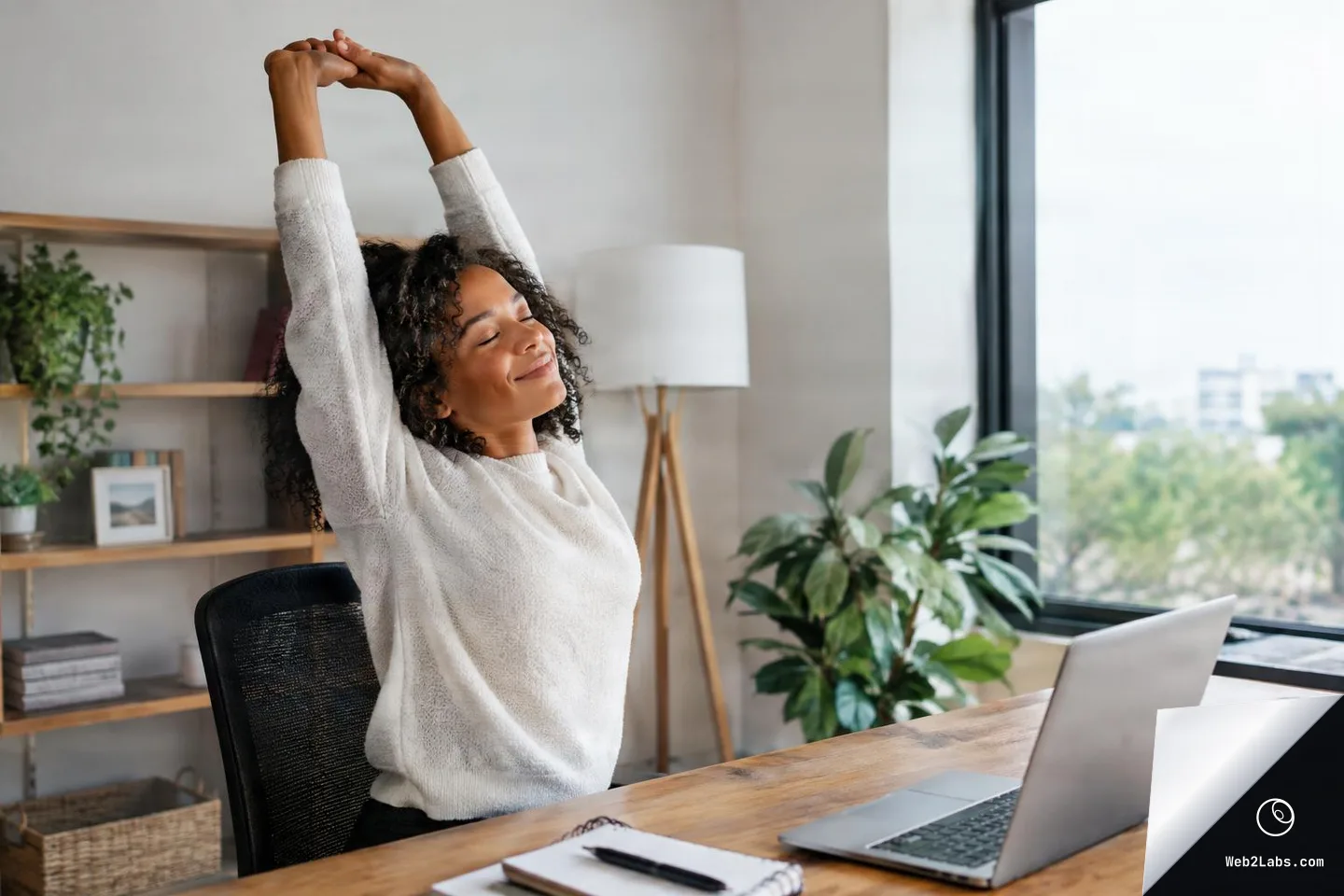 Woman in a white sweater stretches her arms overhead while sitting at a desk with an open laptop and coffee mug in a bright home office by a large window.