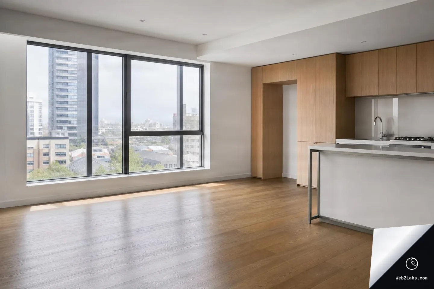 Empty modern apartment interior with wood floors, large black-framed windows showing city buildings, and a compact kitchen with light wood cabinets and a white island.