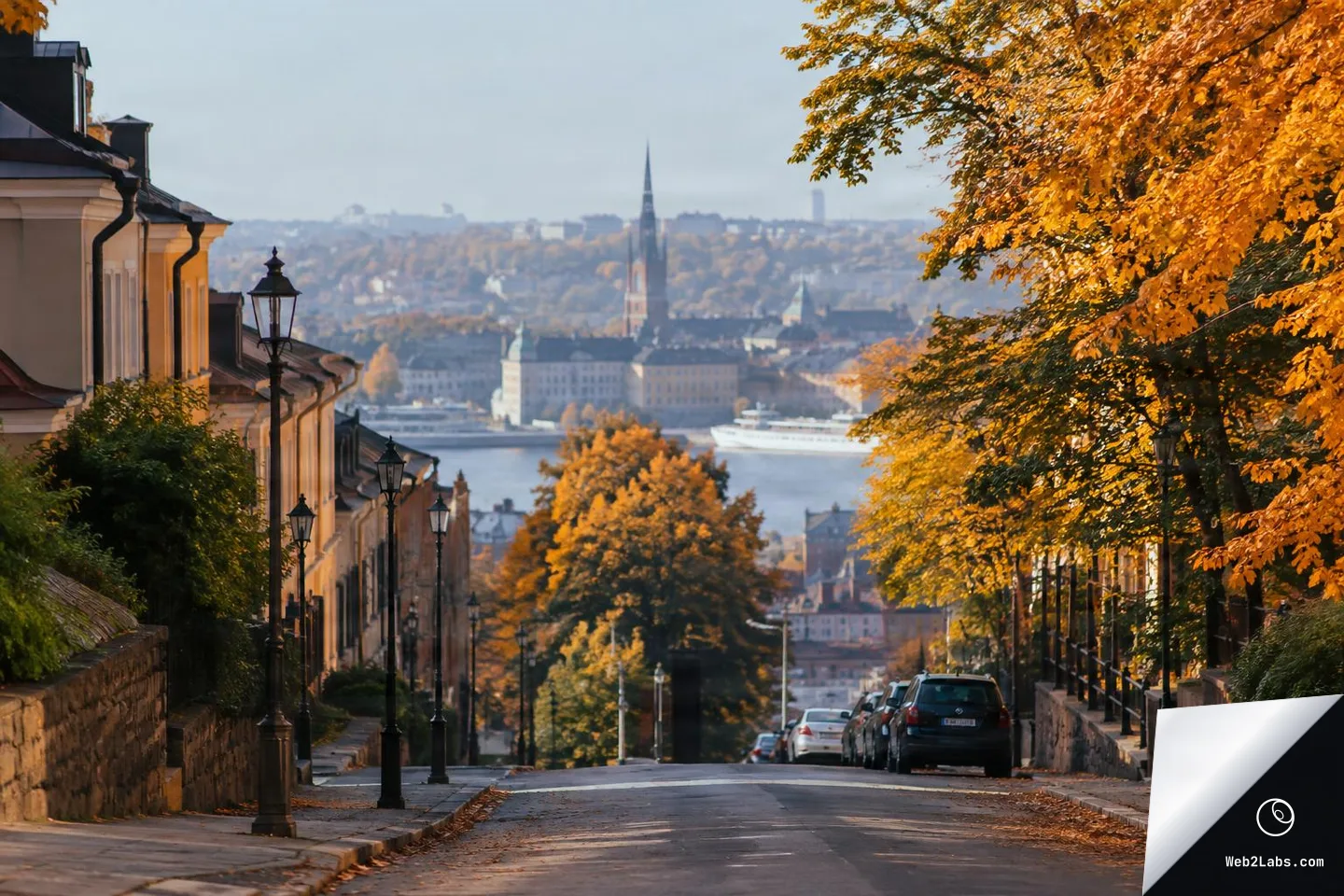 A downhill city street with parked cars and vintage lampposts, framed by bright orange autumn trees, looking toward a distant skyline and waterfront in soft haze.