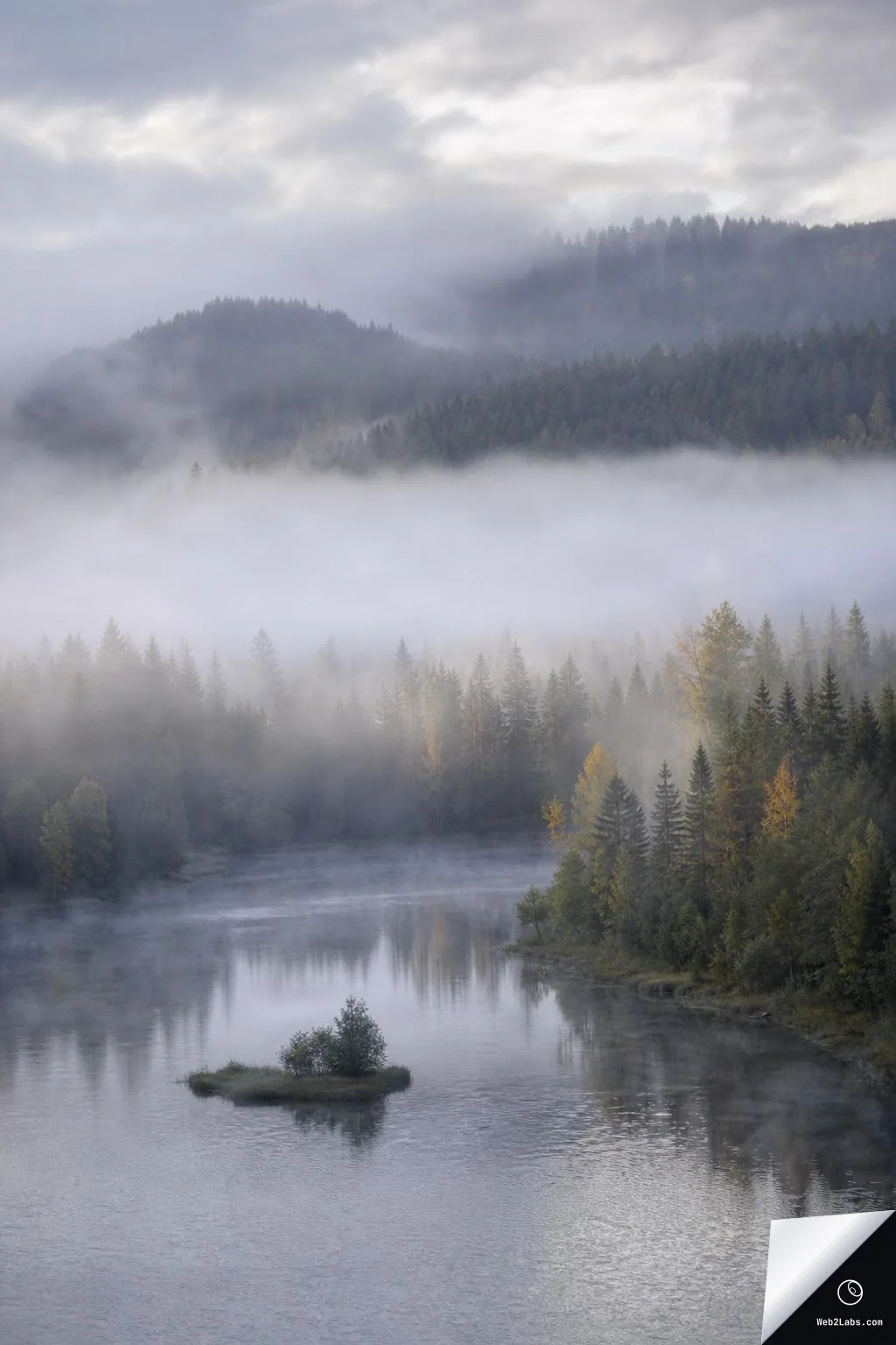 Vertical photo of a calm river with a small shrub-covered islet, bordered by dense evergreen forest and autumn-tinted trees, with thick fog drifting through the valley and hills under a cloudy sky.