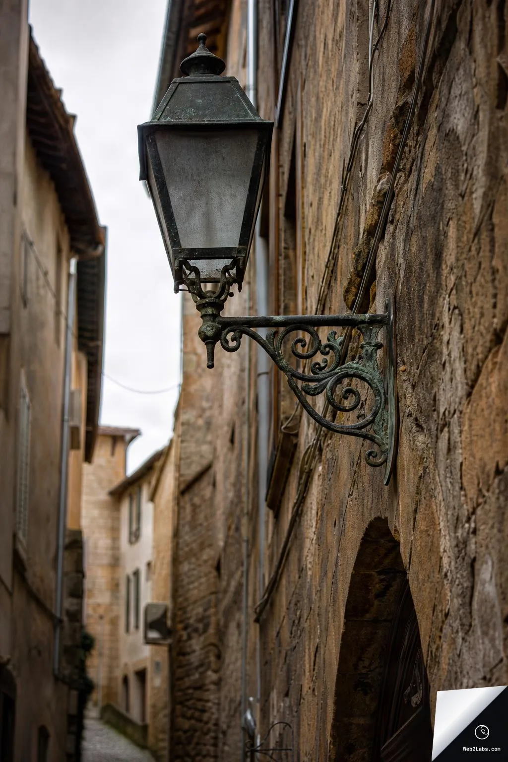 Close-up of a vintage metal street lantern on an ornate bracket attached to a rough stone wall, with a narrow old town alley and buildings blurred in the background.