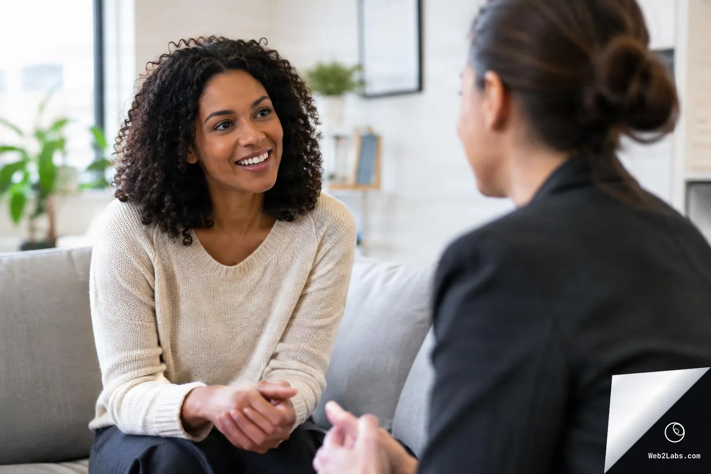 Smiling woman sitting on a couch talking with a business professional in a bright office, with the other person blurred in the foreground