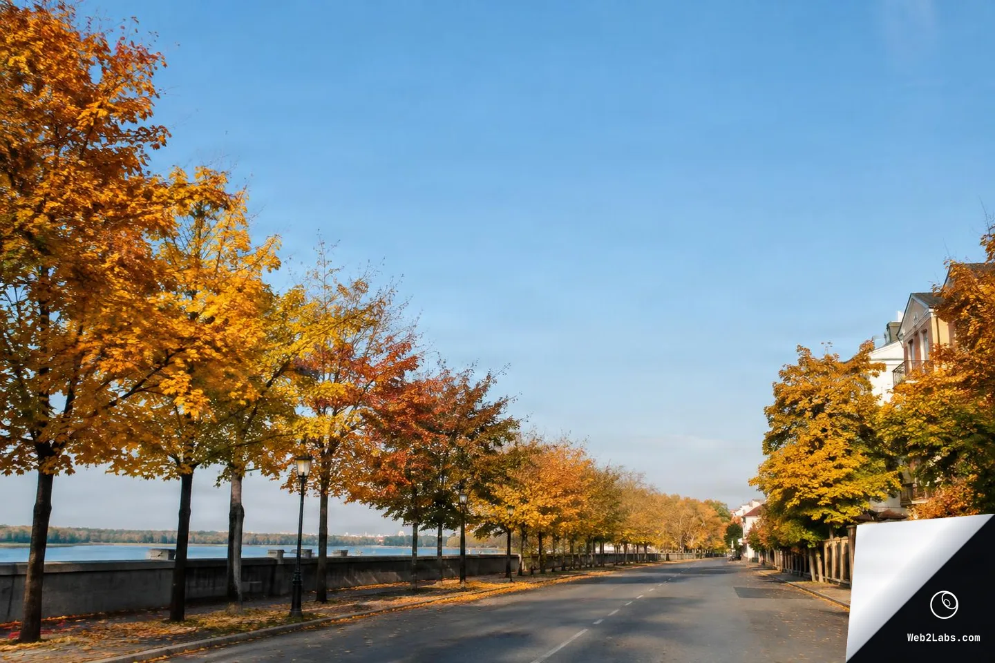 Empty riverside road lined with orange and yellow autumn trees, a stone embankment and water on the left, buildings on the right, and a large clear blue sky above.