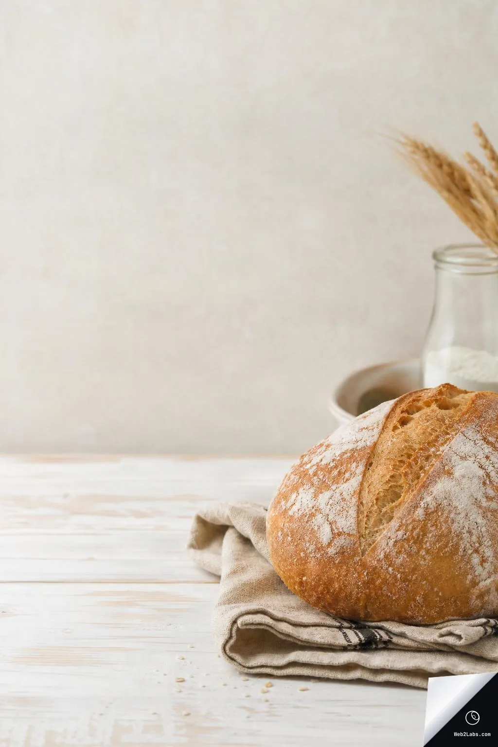 Crusty round loaf of bread on a folded linen cloth on a white wooden table, with a neutral beige background and empty space on the left; a jar of flour and wheat stems are partially visible in the back right.