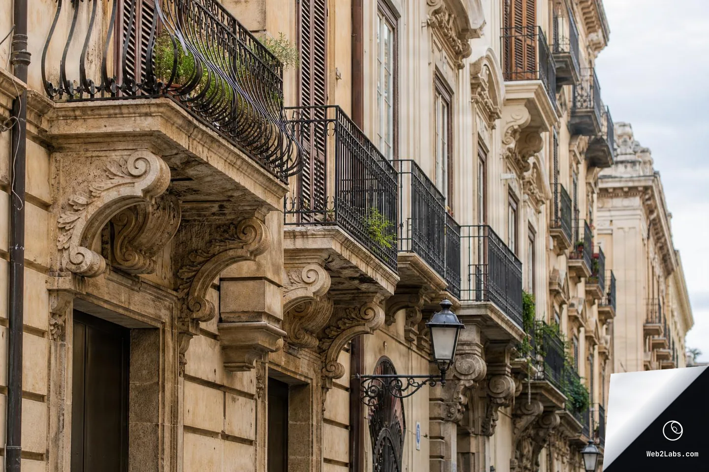 Angled view along historic stone building facades with carved brackets, wrought-iron balconies, shuttered windows, and wall-mounted street lamps under a lightly cloudy sky.
