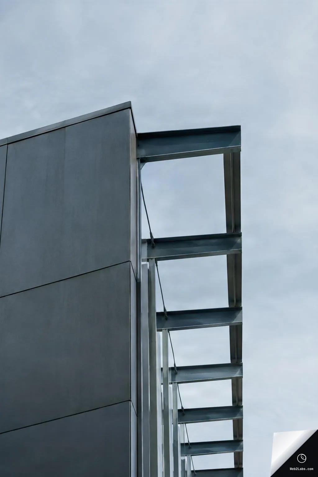 Close-up of a modern building corner with dark metal facade panels and an exposed steel frame structure, set against a cloudy gray sky in a vertical composition.