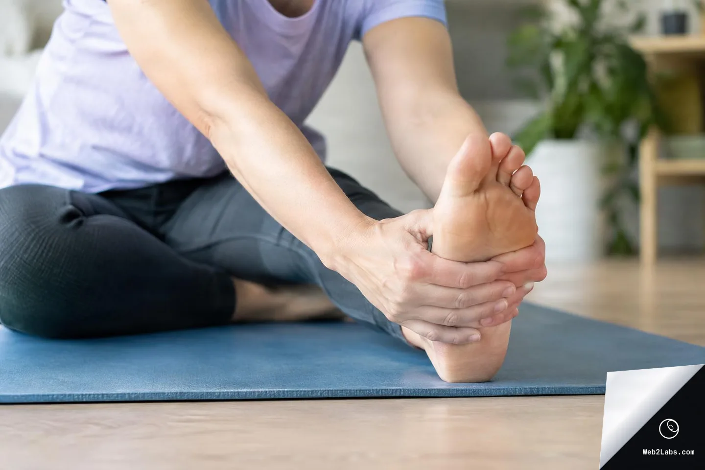 Close-up of a person sitting on a yoga mat and holding their foot while stretching their leg indoors, with a blurred plant in the background.