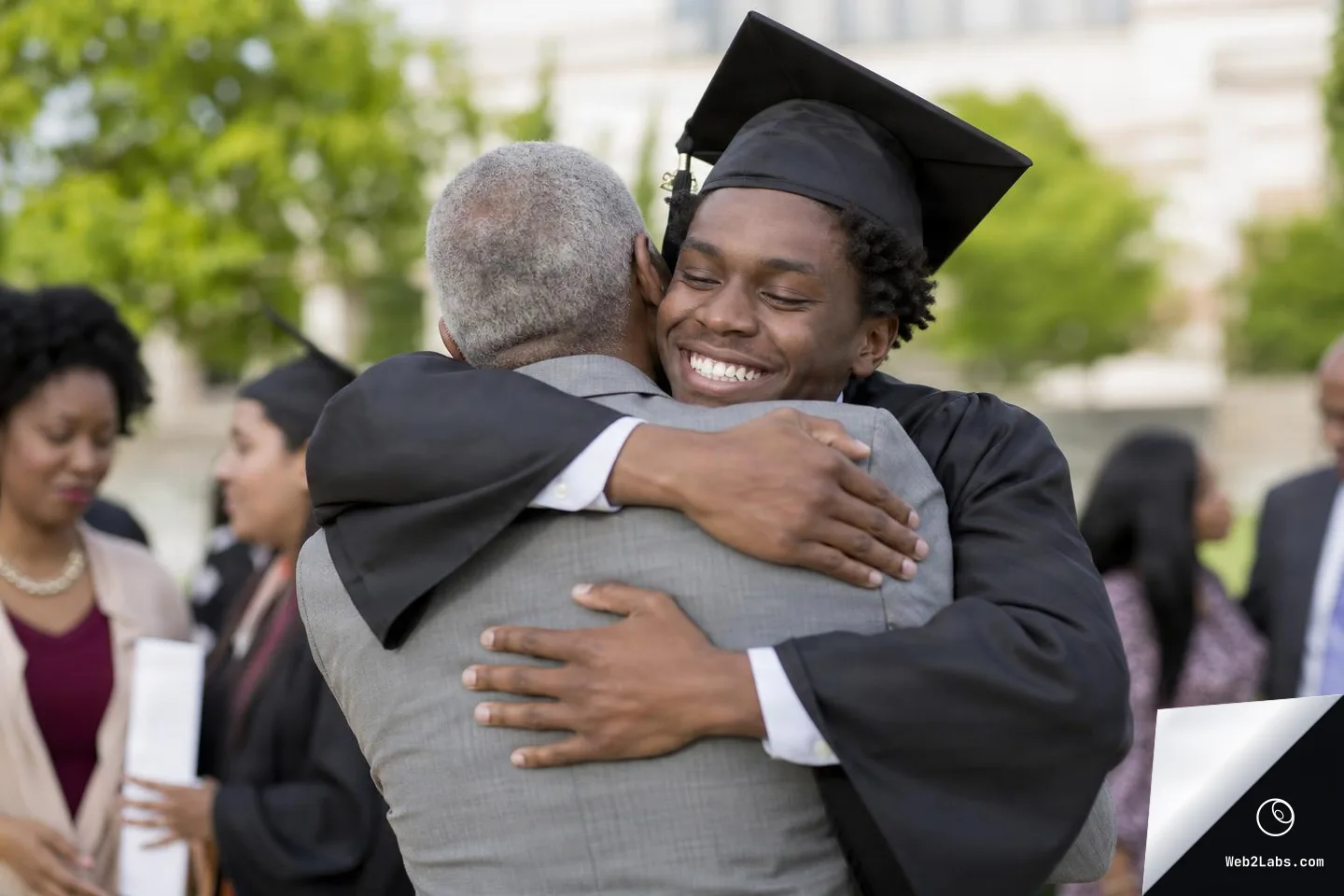 Smiling graduate wearing a black cap and gown hugs an older man in a gray suit outdoors during a graduation gathering, with other people blurred in the background.