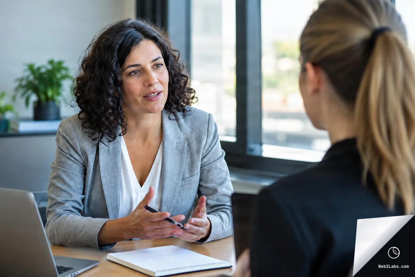 A woman in a gray blazer talks and gestures while holding a pen during a mentoring meeting across a desk, seen over the shoulder of a colleague in a bright office with windows.
