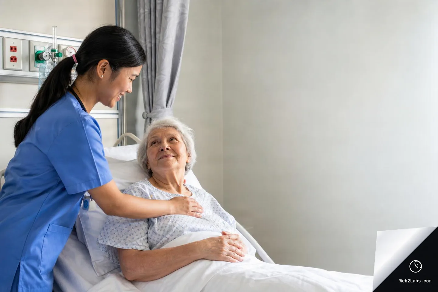 A nurse in blue scrubs smiles while gently touching the shoulder of an elderly woman patient resting in a hospital bed, with medical wall outlets behind them and open blank wall space on the right.