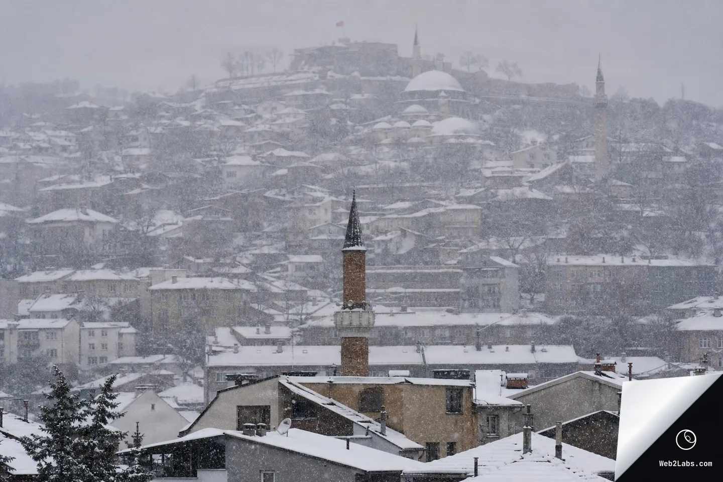 Telephoto view of a hillside town during heavy snowfall, with snow-covered rooftops in the foreground and layered buildings fading into a gray, snowy background; a tall tower rises near the center.