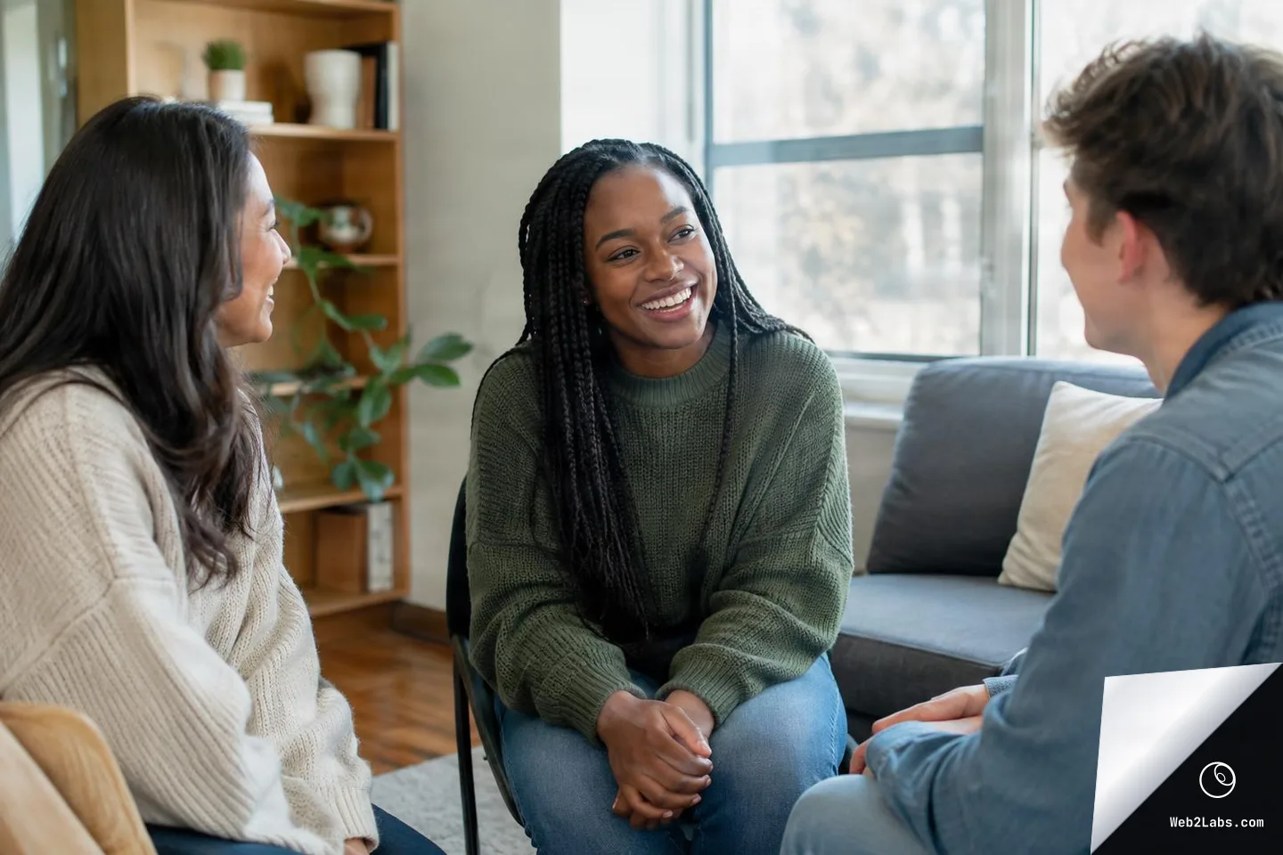 Three young adults sit in a small circle in a bright living room, smiling and talking supportively, with a sofa and bookshelf in the background.