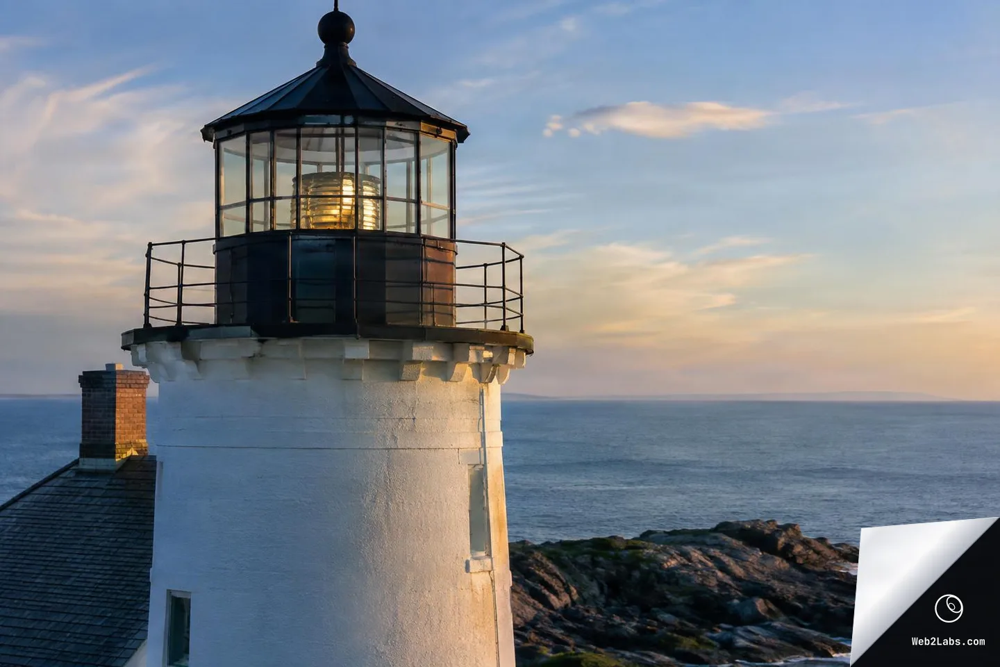 Close-up of the top section of a white coastal lighthouse with a black lantern room and glowing lens, overlooking a calm ocean and rocky shoreline at sunset.