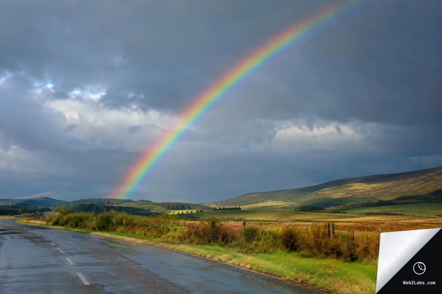 Wide landscape photo of a vivid rainbow arcing through dark storm clouds above green and brown hills, with a wet rural road and roadside fence in the foreground.