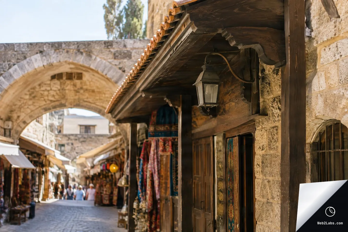 Close-up of a wooden shop awning with a metal lantern mounted on a stone wall, looking down a cobblestone market lane toward a large stone arch and stalls in the distance.