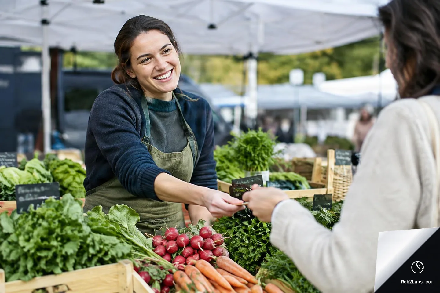 Smiling market stall vendor in an apron hands an item to a customer over a display of radishes, carrots, and leafy greens at an outdoor farmers market under a canopy.