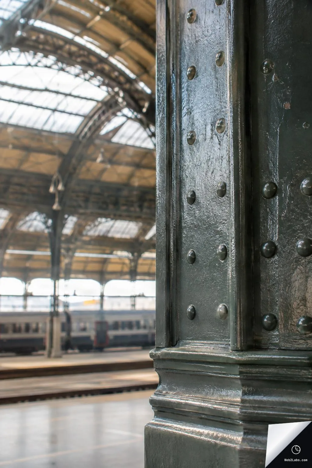 Close-up of a dark painted riveted steel column in a railway station hall, with the arched glass-and-steel roof and a blurred train platform in the background.