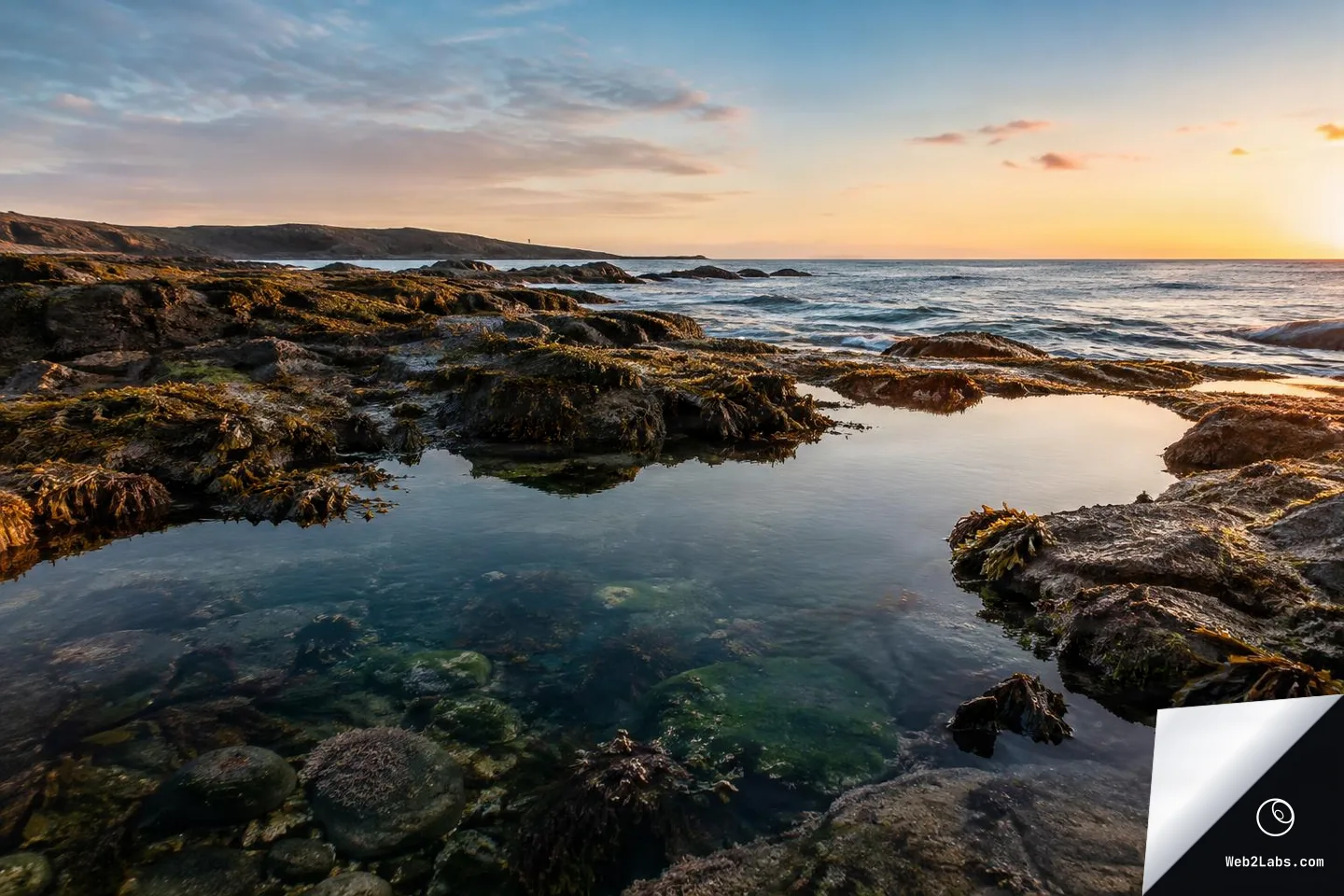 Wide view of a rocky shoreline at sunset with a clear tidal pool in the foreground, seaweed-covered rocks around it, and the ocean and distant headland under a partly cloudy sky.
