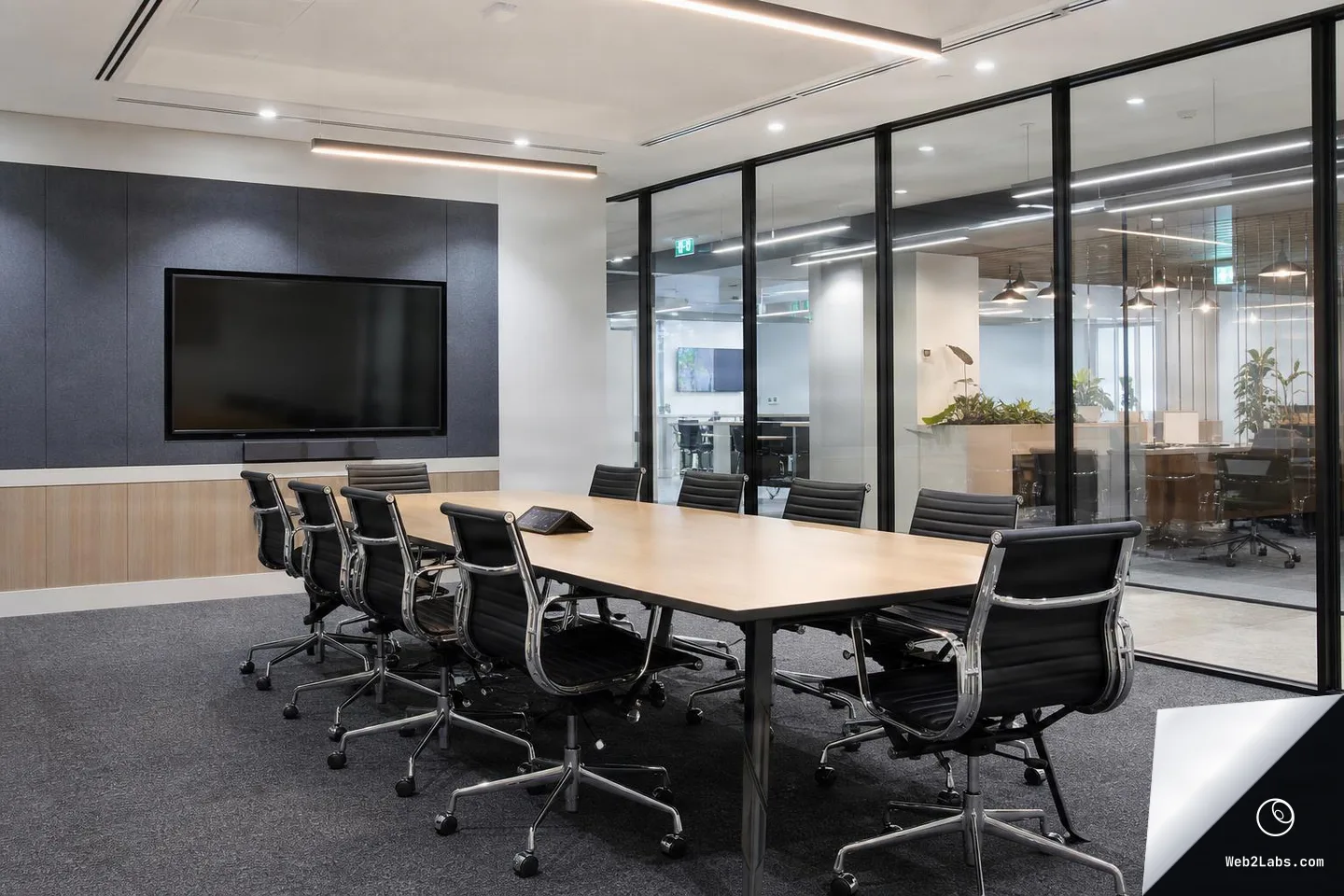 Empty modern conference room with a long wooden table, black office chairs, a wall-mounted TV screen, and glass walls looking into an open-plan office area.