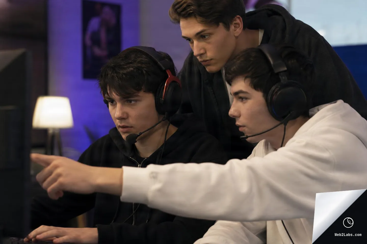 Three young men wearing gaming headsets collaborate at a computer; one points at an off-screen monitor while the others watch closely in a softly lit room.