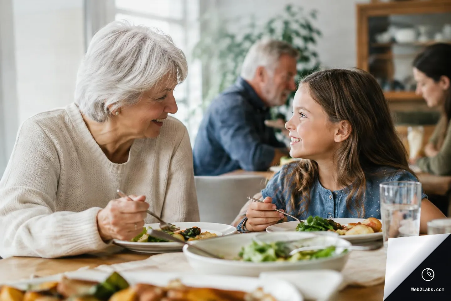 An elderly woman and a young girl sit at a dining table with plates of food, smiling at each other while holding forks; other family members are blurred in the background.