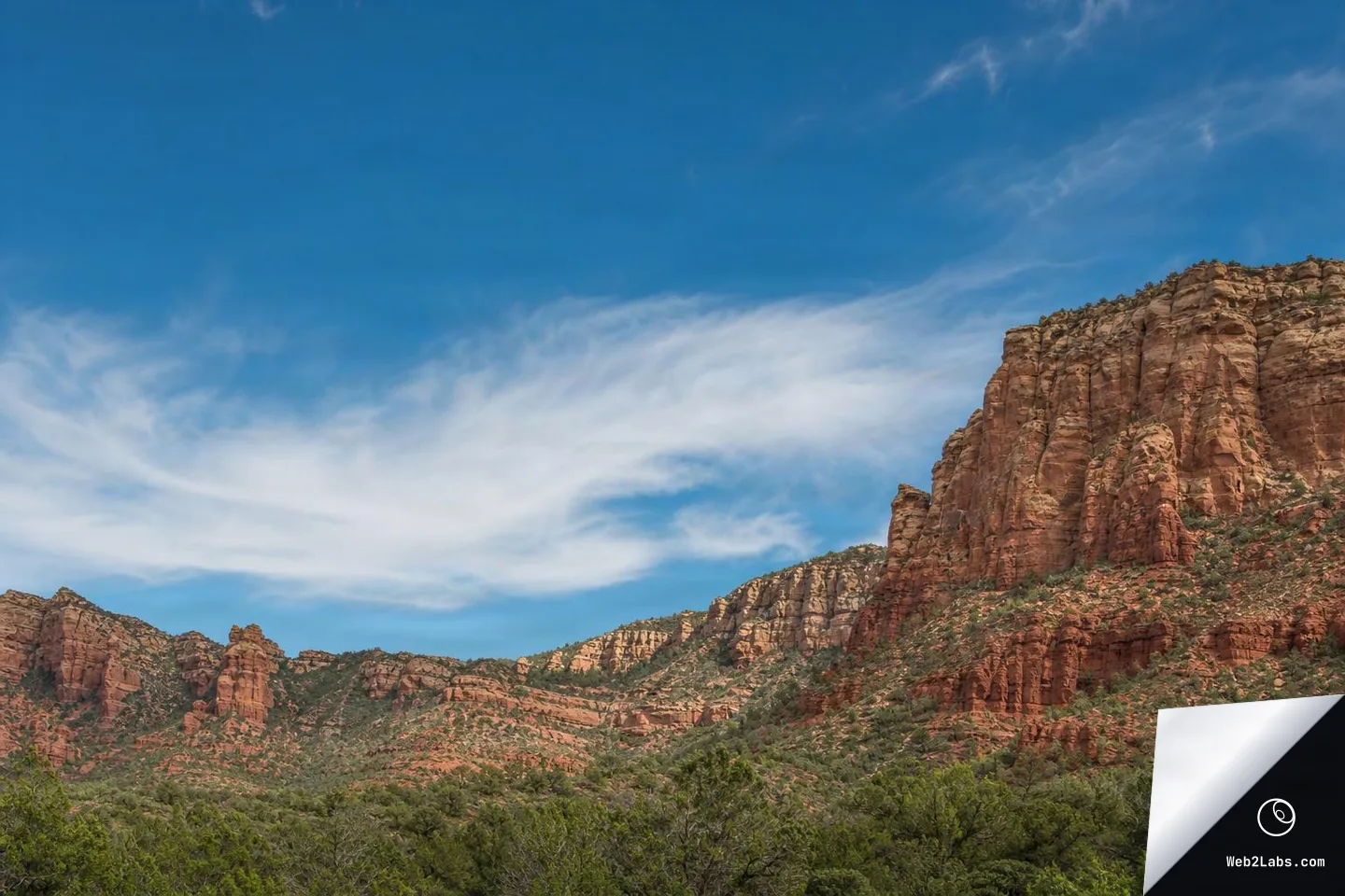 Red rock canyon cliffs and green scrubland beneath a wide blue sky with thin white clouds, framed as a horizontal landscape with large open sky space.