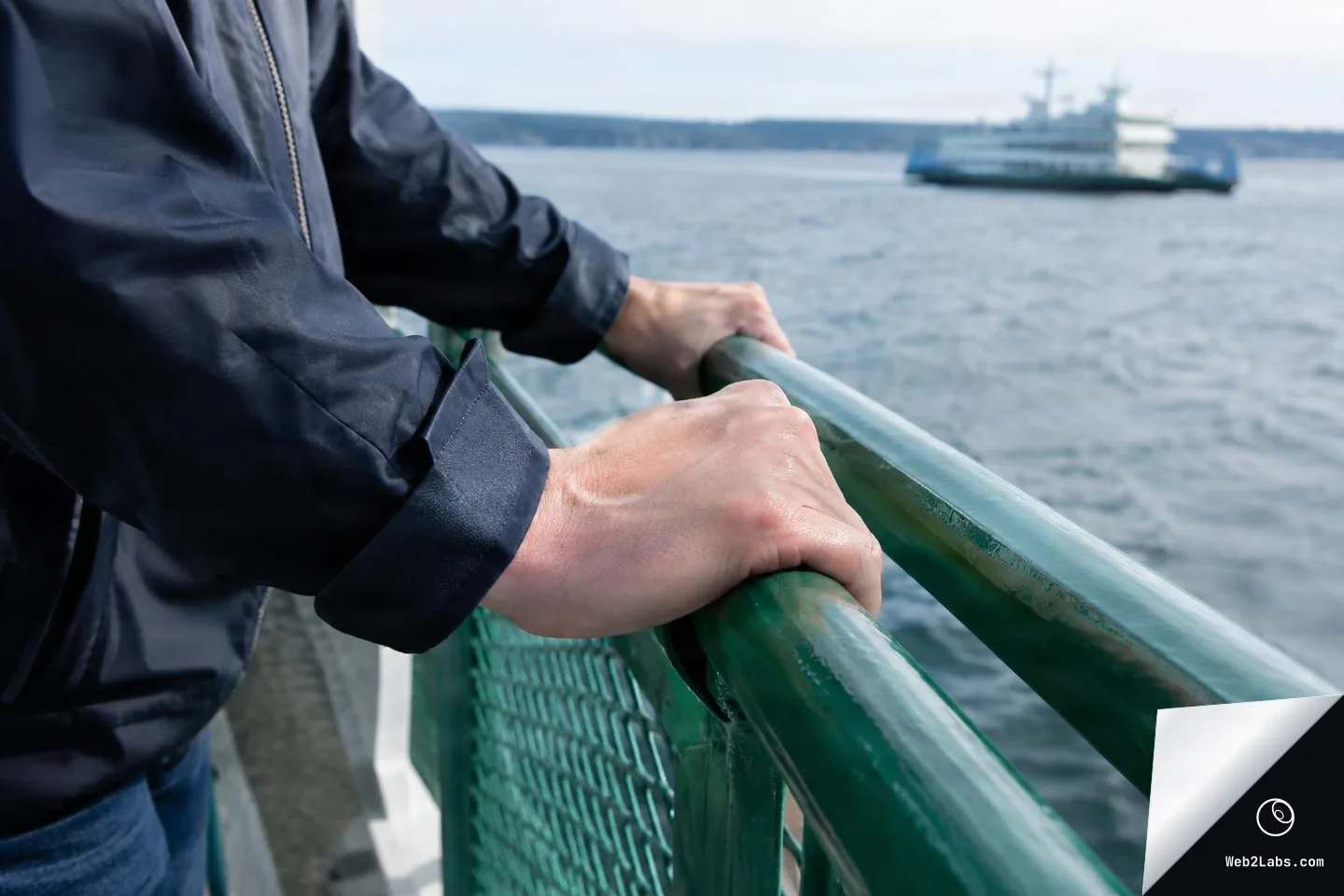 Close view of a person in a dark jacket gripping a green railing on a ferry, with water and a blurred ferry boat in the distance.
