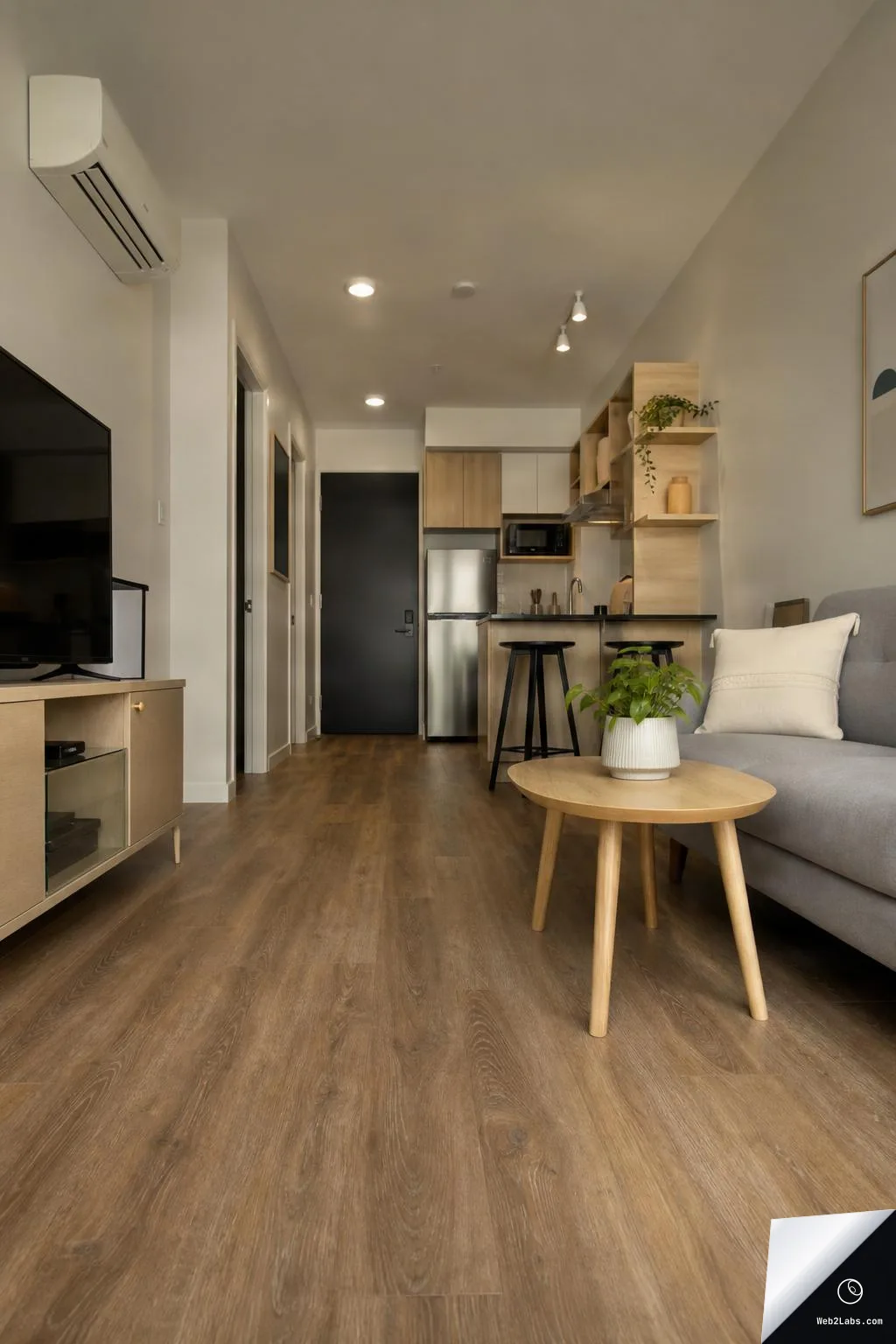 Low-angle view down a small modern studio apartment with wood floors, a grey sofa and round coffee table with a potted plant, and a compact kitchen with bar stools and open shelving in the background.