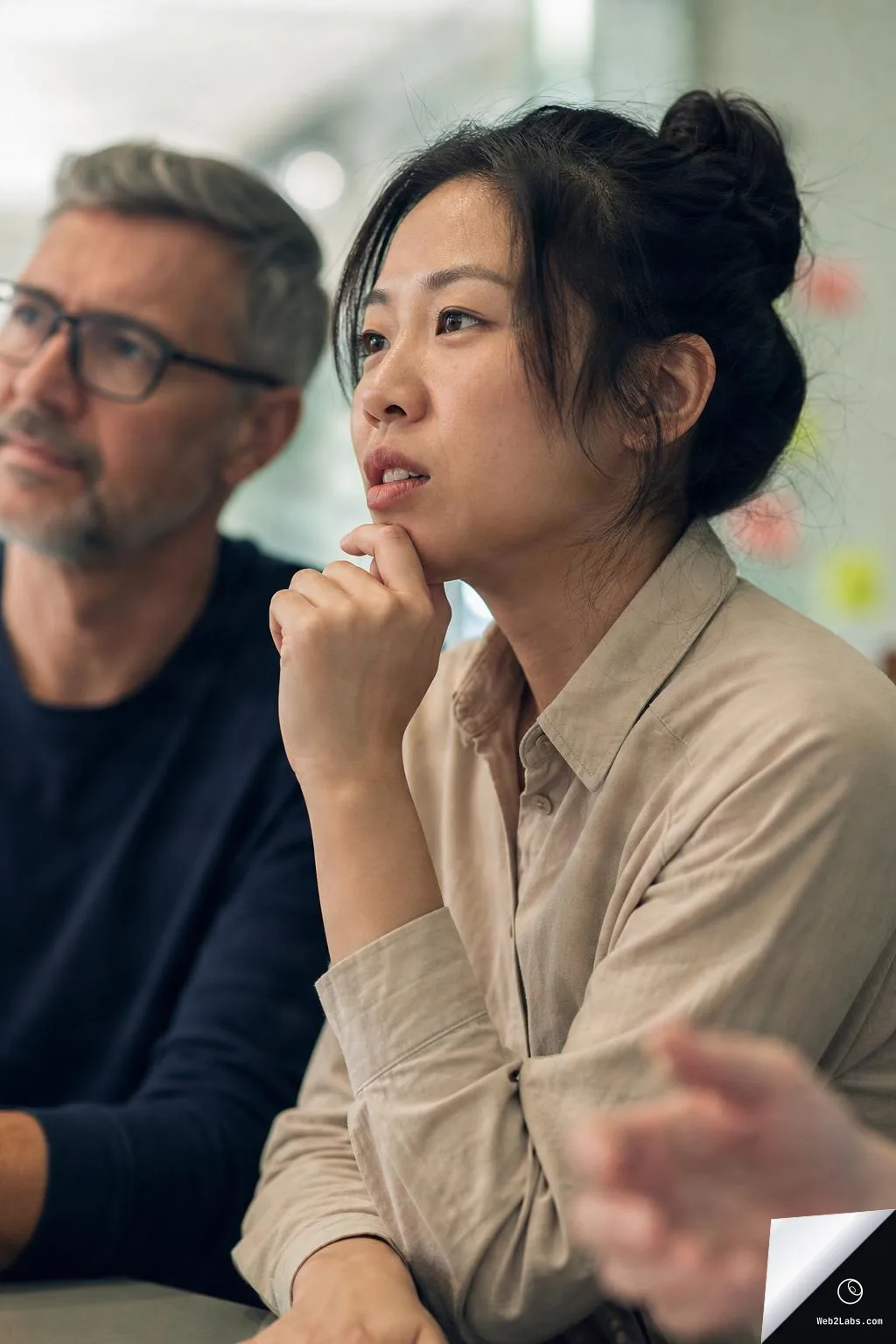 Close-up of a thoughtful woman with her hand on her chin in a meeting, with a man in glasses blurred in the background and an out-of-focus hand gesturing in the foreground.