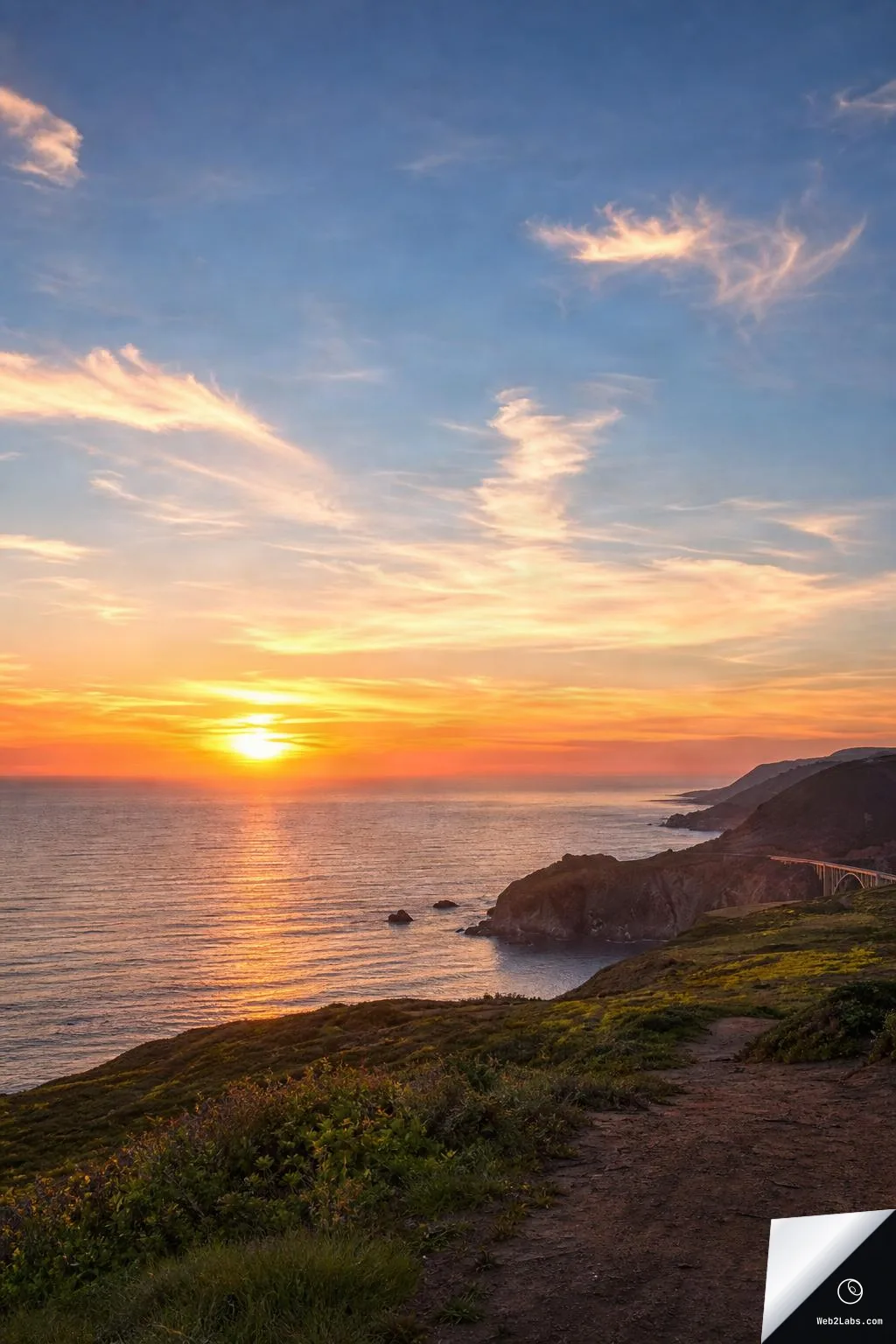 Sun setting over a calm ocean with orange reflections, wispy clouds in a blue sky, and dark coastal cliffs with a grassy hillside in the foreground.