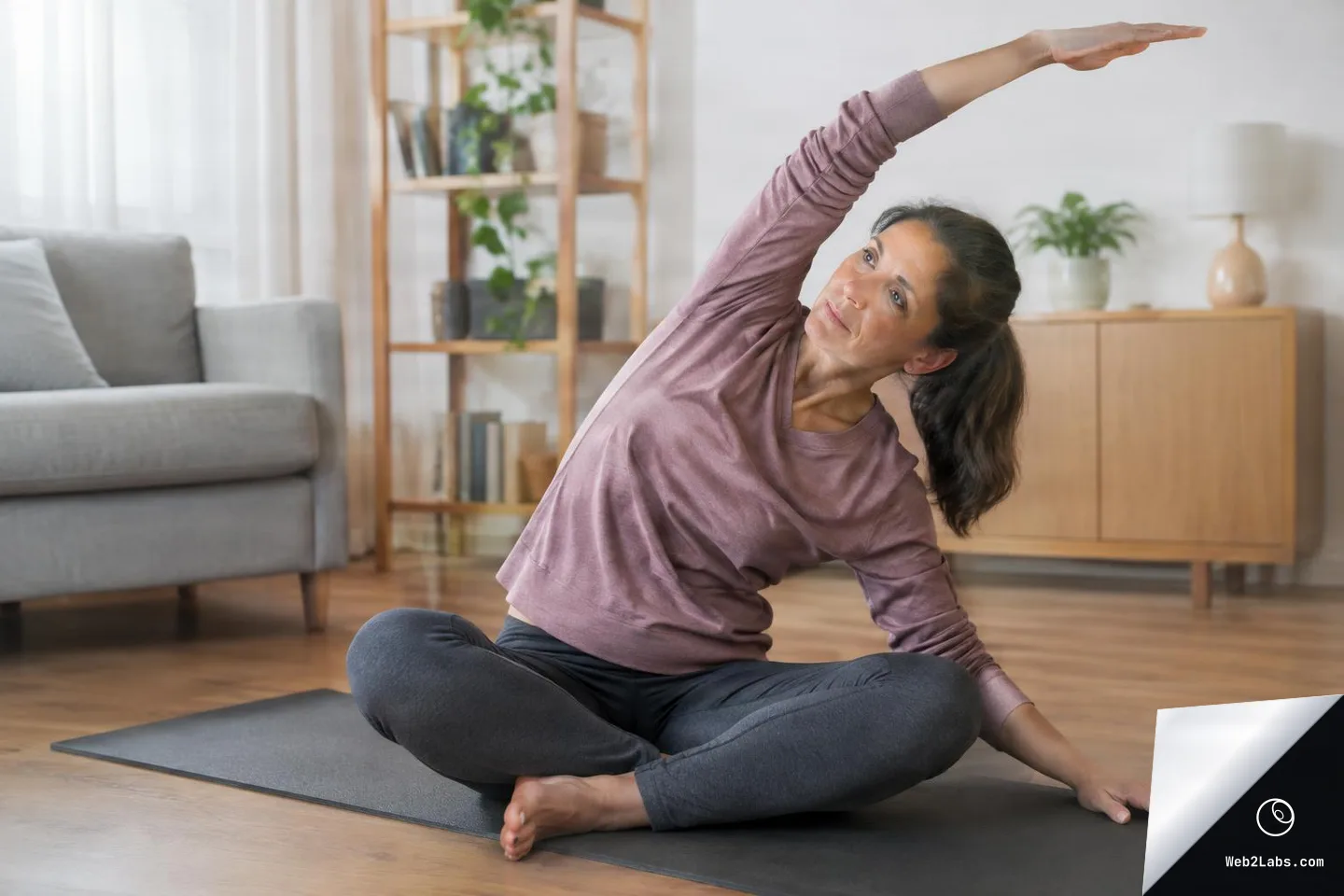 Woman seated on a yoga mat in a living room performing a side stretch with one arm reaching overhead.