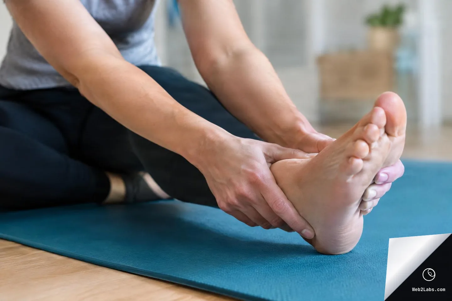 Close-up of a person seated on a blue yoga mat, holding their foot with both hands while stretching the leg indoors.