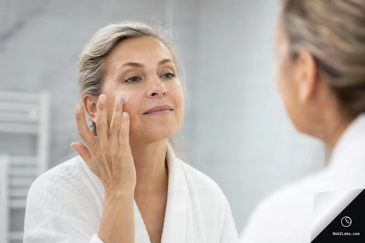 Mature woman in a white bathrobe applying face cream to her cheek while looking at herself in a bathroom mirror, with her reflection softly blurred in the foreground.