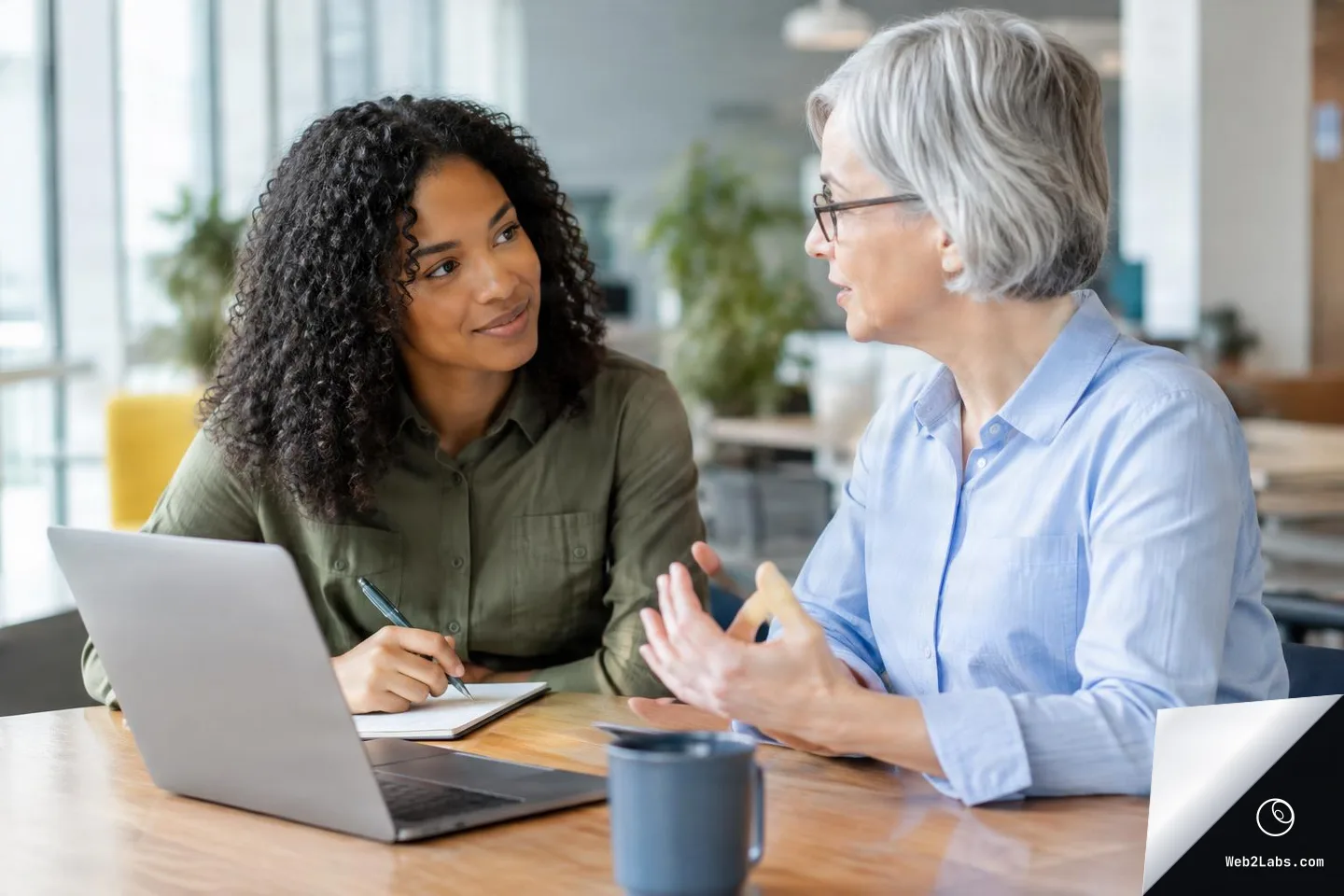 Two coworkers in a bright office sit at a wooden table, making eye contact while one gestures and the other takes notes next to an open laptop.